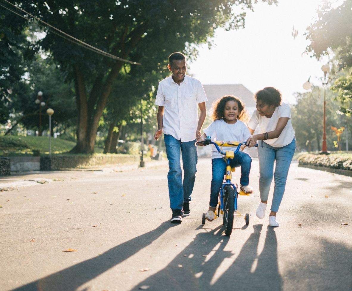 Black mom and dad teaching their daughter how to ride a bike in the street.